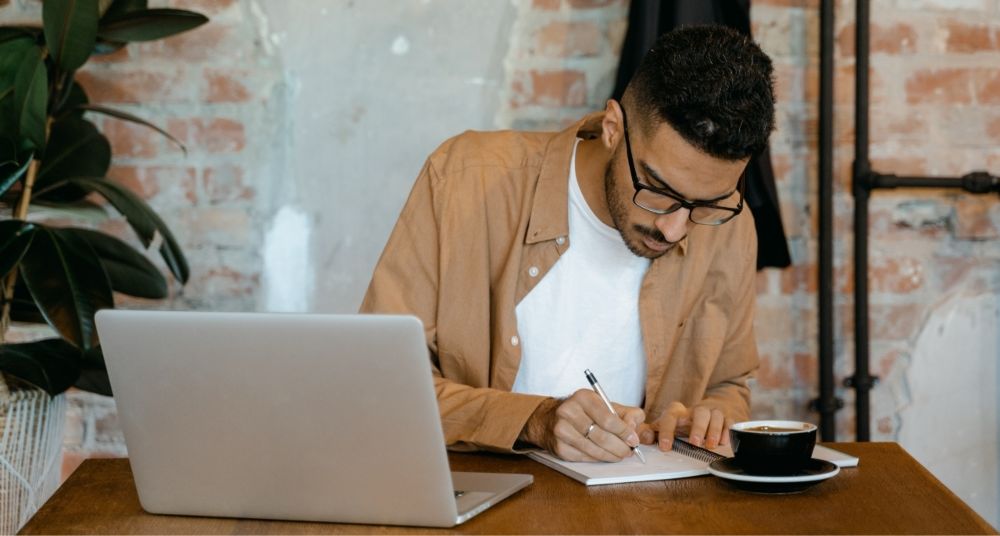 A man wearing glasses and a brown shirt is writing on a notebook.