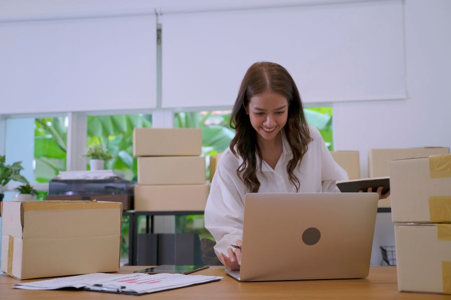 Online seller working at a laptop surrounded by packing boxes in a small workspace.