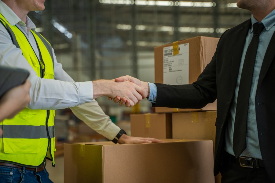 Warehouse manager and hi-vis worker shaking hands amid stacked cardboard boxes.