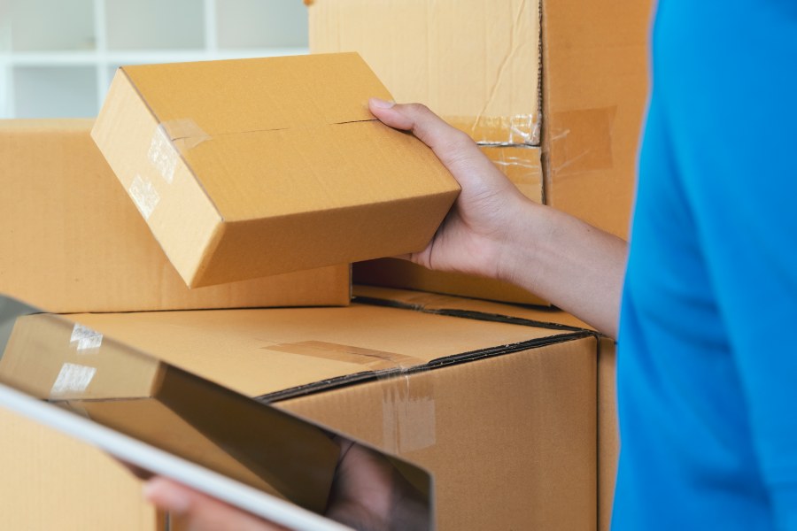 Worker inspecting taped cardboard box while checking tablet inventory list in warehouse.