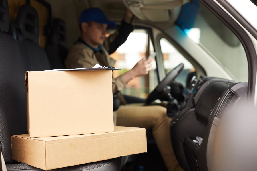 Delivery driver in van cabin holding stacked cardboard parcels, clipboard and phone.