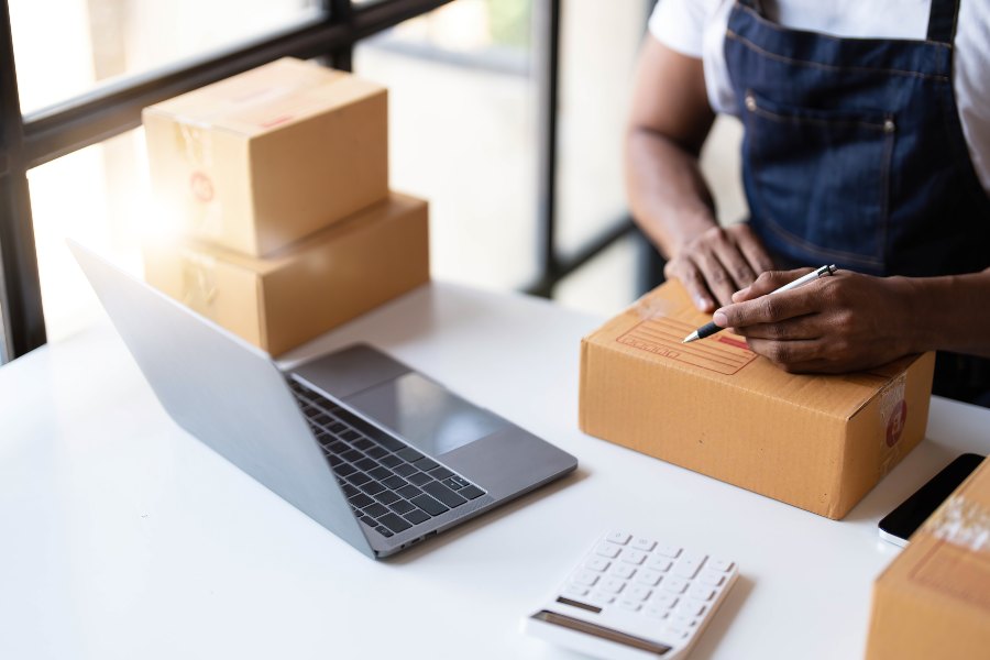 Entrepreneur in apron labeling cardboard box beside laptop and calculator for e-commerce shipping.