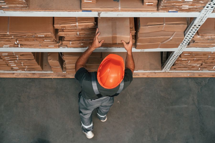 Warehouse worker in orange hard hat lifts cardboard box from shelf, focusing on accurate order picking.