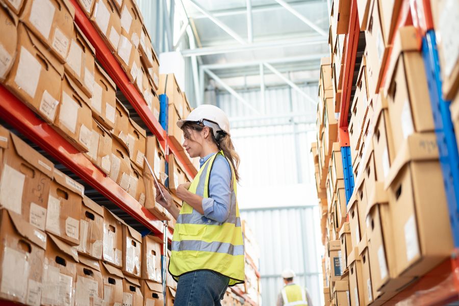 Warehouse worker in hi-vis vest and hard hat checks labelled cartons on racks for accurate order picking.