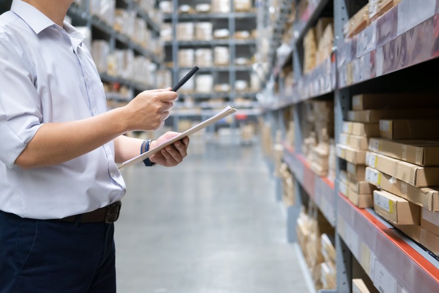 Man in white shirt holds tablet, scanning goods amid warehouse shelves and boxes.
