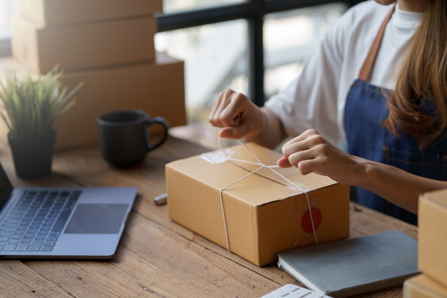 Woman ties string around shipping box on desk with laptop and plants.