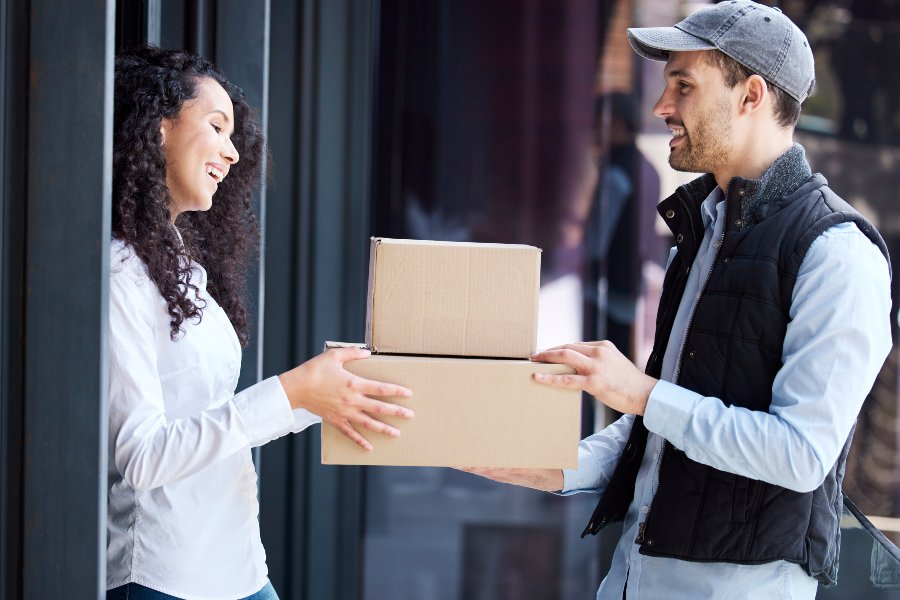 Smiling delivery man hands two boxes to woman at building entrance.