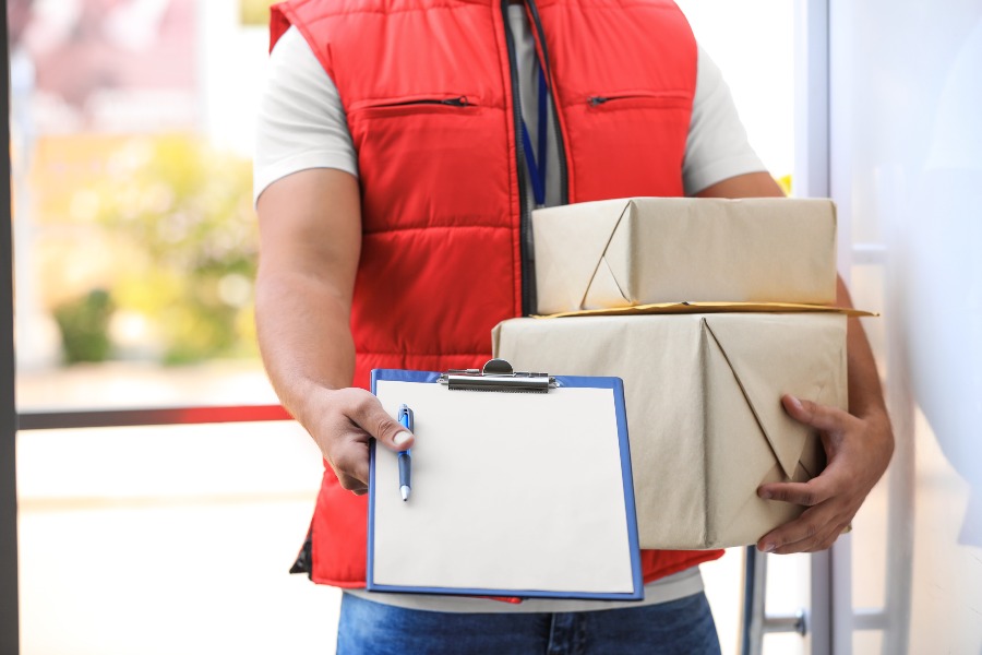 Courier in red vest holds stacked packages and clipboard at doorway entrance.