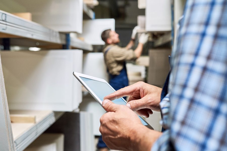 Warehouse worker uses tablet for inventory check as colleague stocks shelves behind.