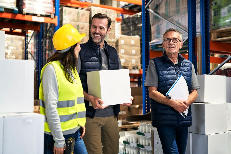 Warehouse team discusses operations while holding boxes in a storage aisle.
