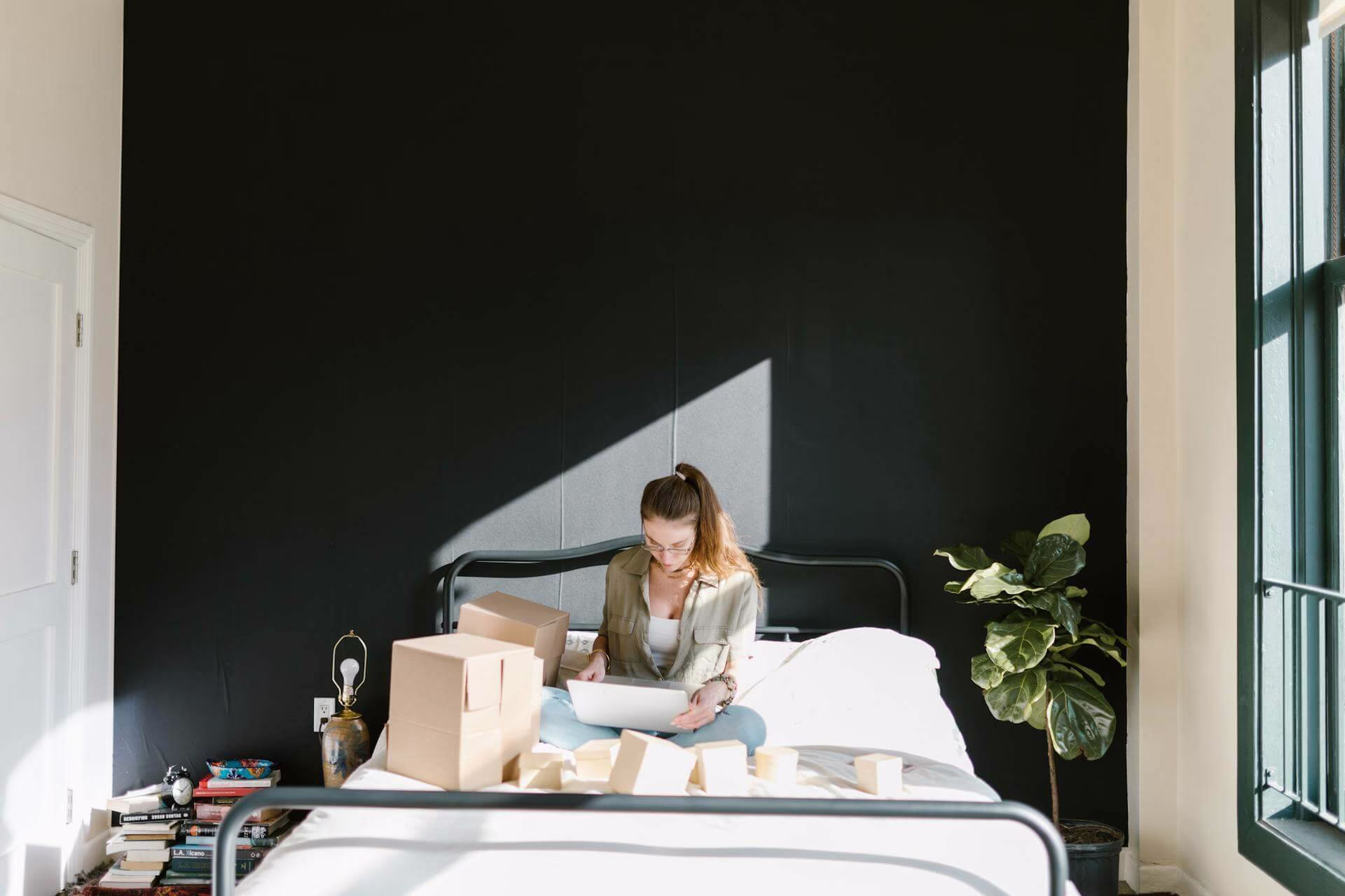 A woman sitting on a bed with boxes of items in front of her.