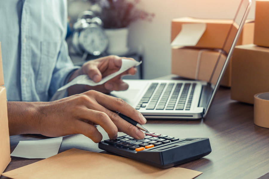 Person using a calculator and laptop beside cardboard boxes to assess inventory costs, including dead stock.
