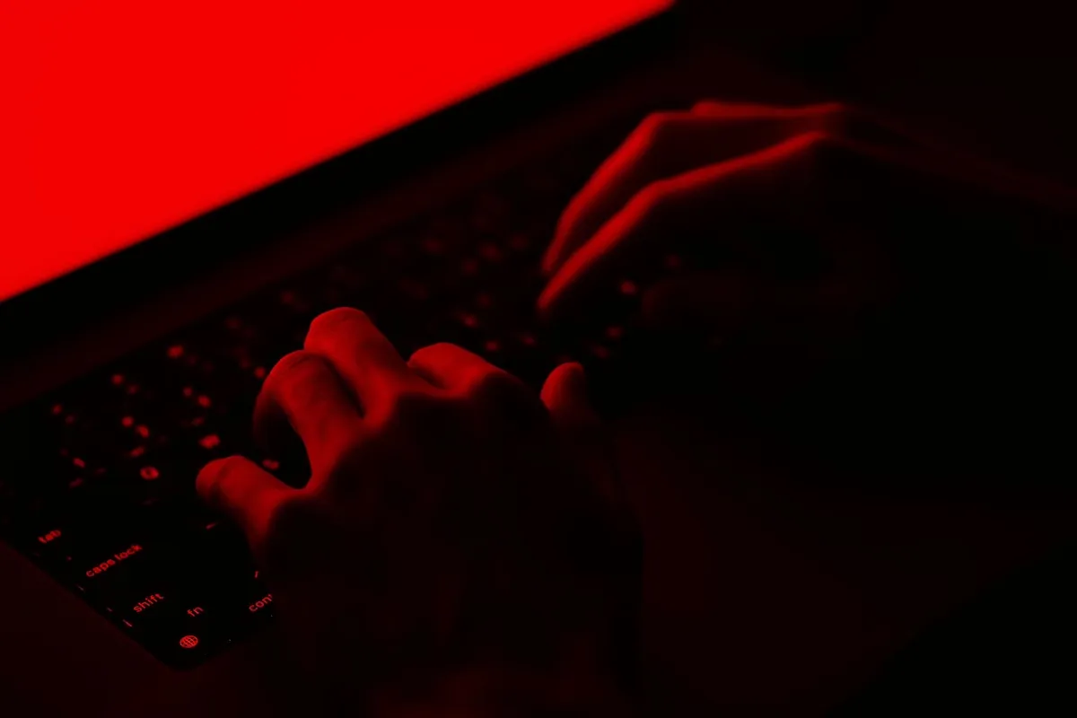 Hands typing on a keyboard illuminated by red light from a screen in a dark setting.