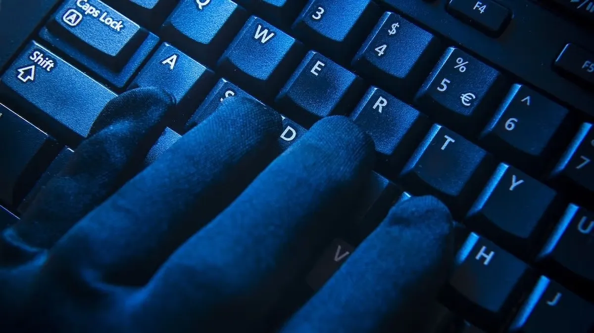 Close-up of fingers wearing black gloves typing on a black computer keyboard with blue backlight.