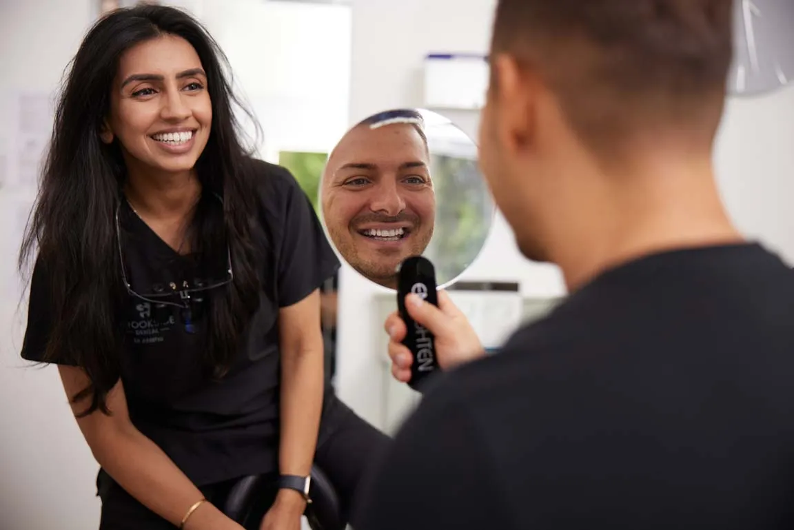 Smiling woman in black scrubs watches a man holding a round mirror reflecting his smiling face.