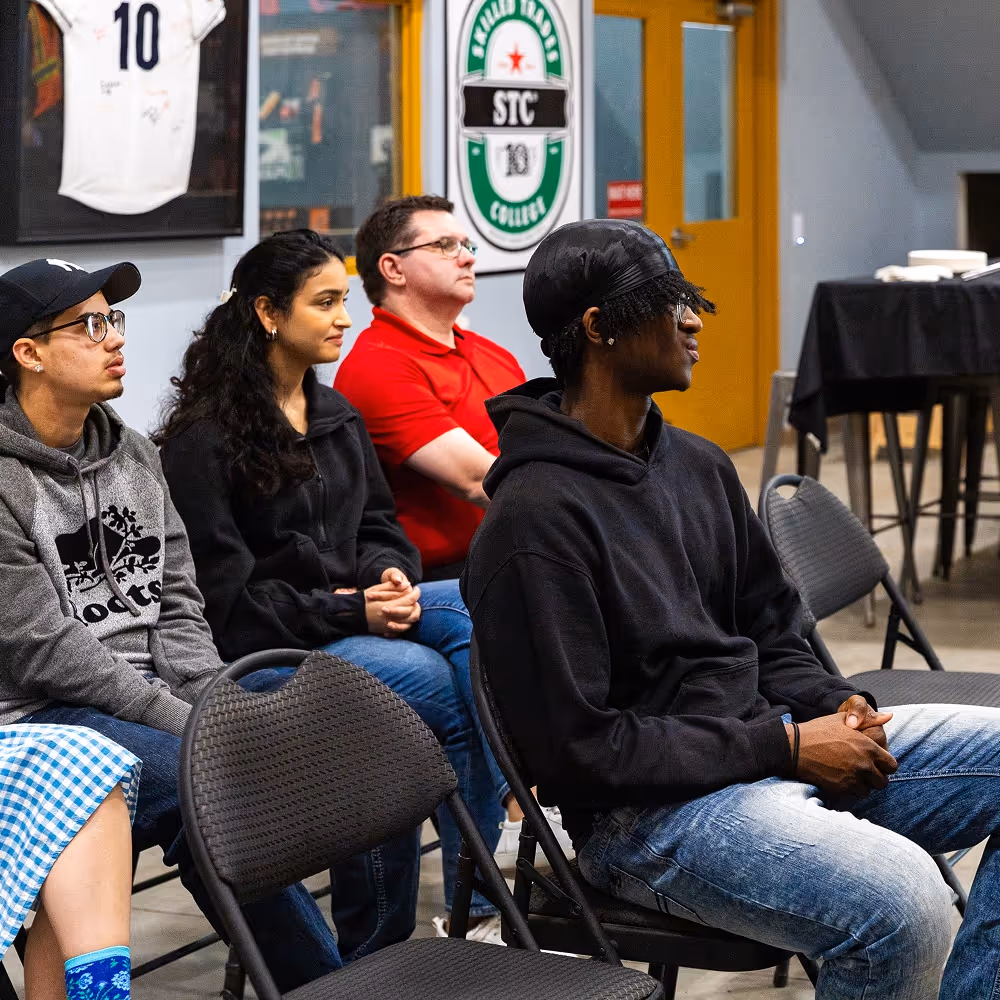 Four people seated in chairs attentively listening in a room with sports memorabilia on the walls.