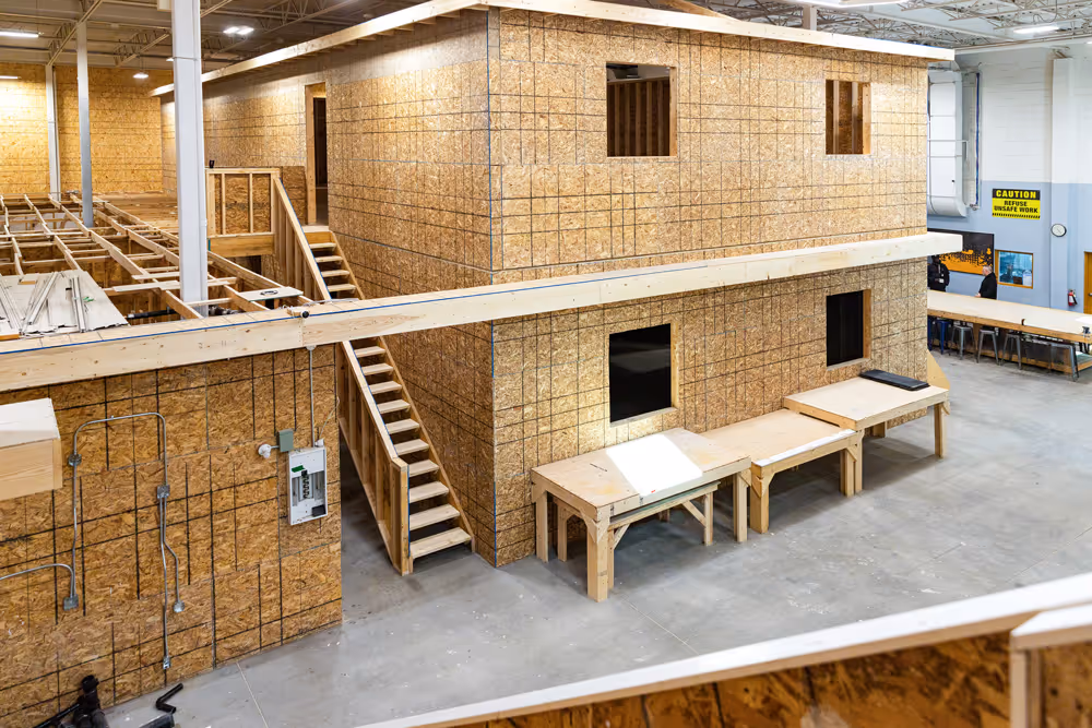 Interior view of a large workshop with a two-story wooden structure under construction, workbenches, and a caution sign in the background.