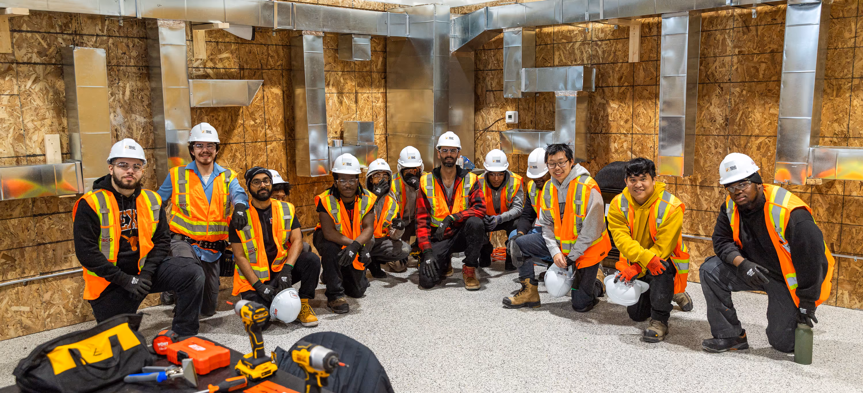 Group of construction workers wearing orange safety vests, gloves, and some with white hard hats, posing and kneeling inside a building under construction with metal ductwork on plywood walls.