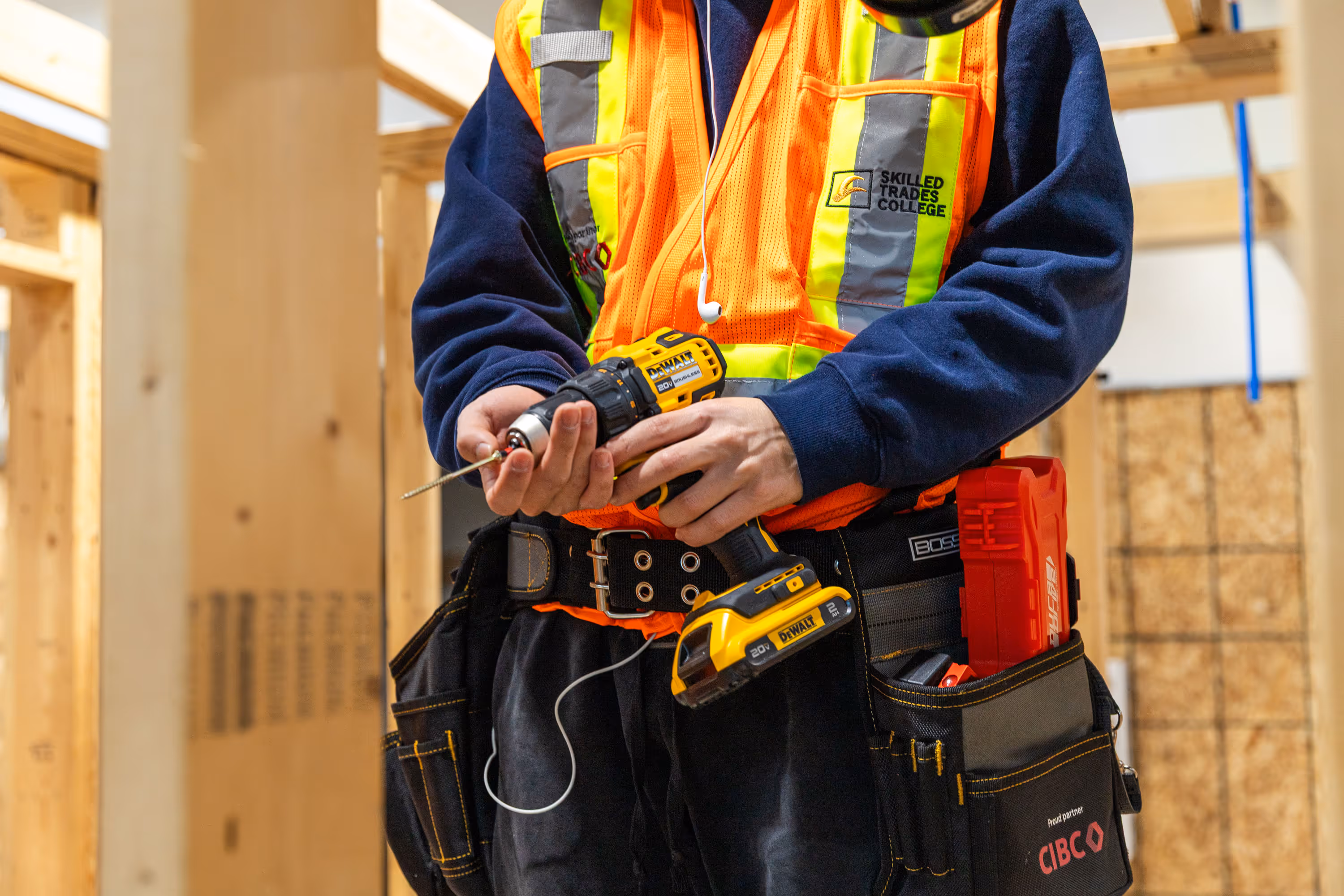 Construction worker wearing a high-visibility vest holding a DeWalt power drill inside a wooden frame building.