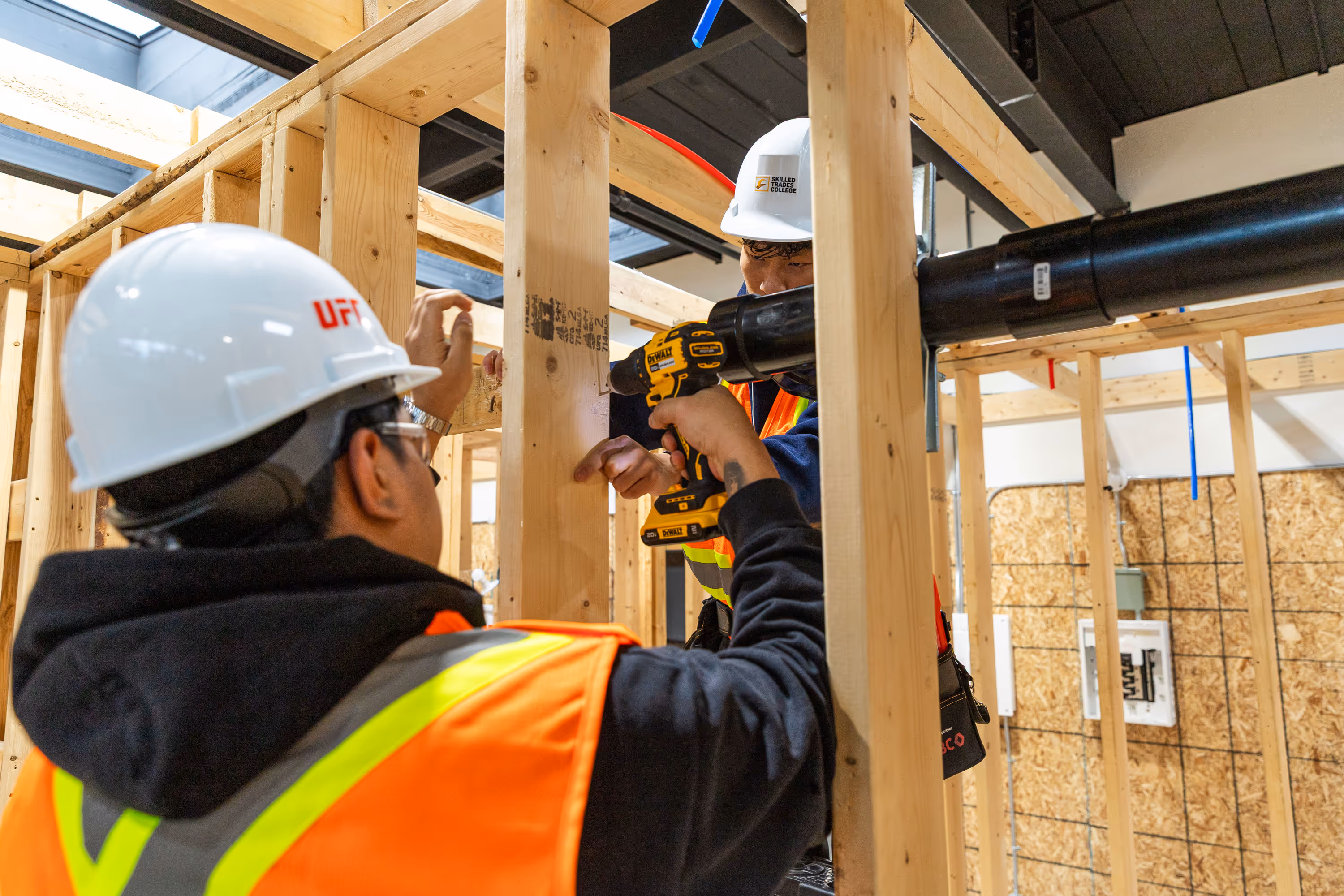 Two construction workers wearing white hard hats and orange safety vests installing a black pipe on a wooden frame using a power drill.