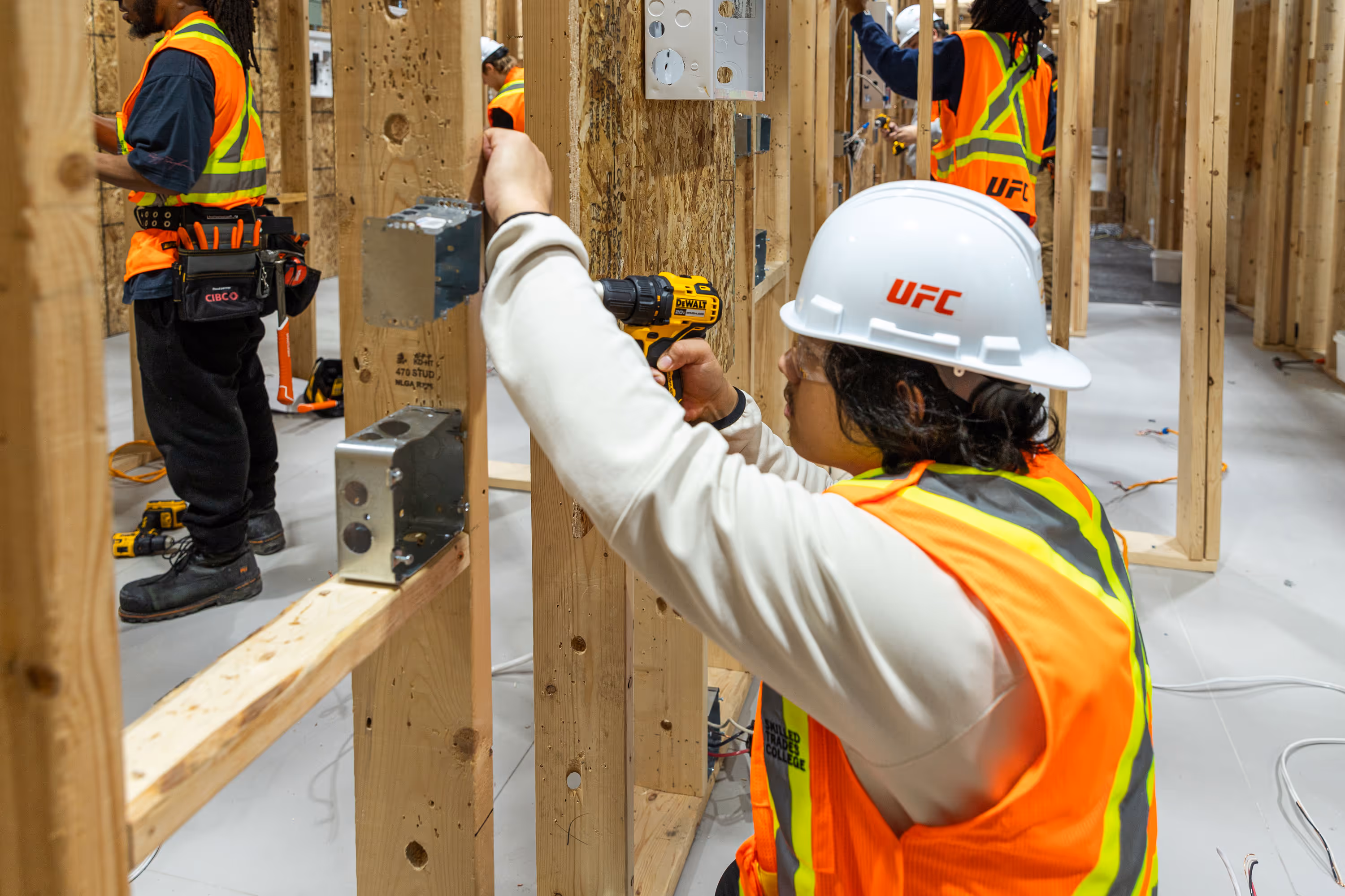 Construction workers in safety vests and helmets installing electrical boxes on wooden studs in a building under construction.