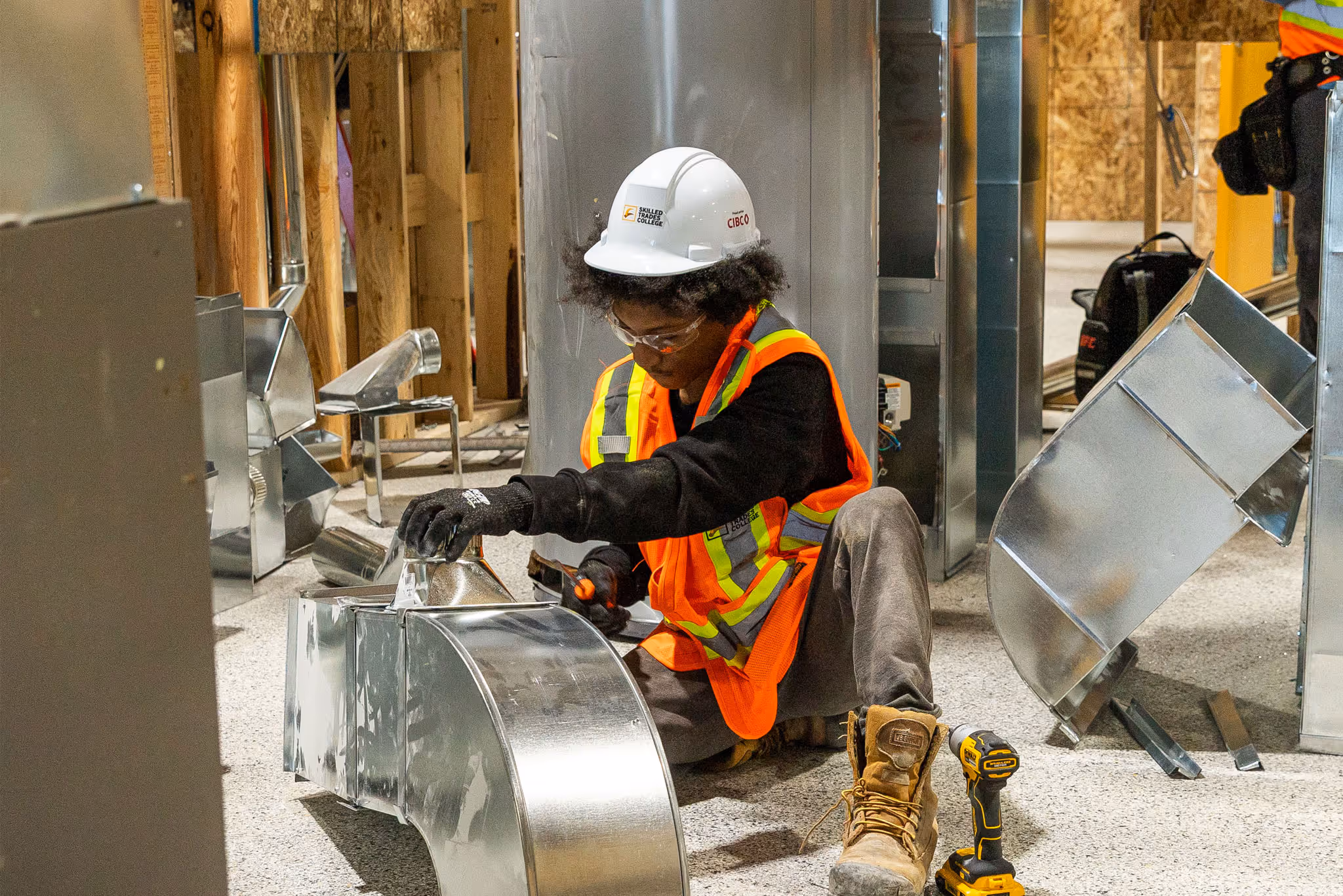 Construction worker wearing a white hard hat and orange safety vest assembling metal ductwork indoors.