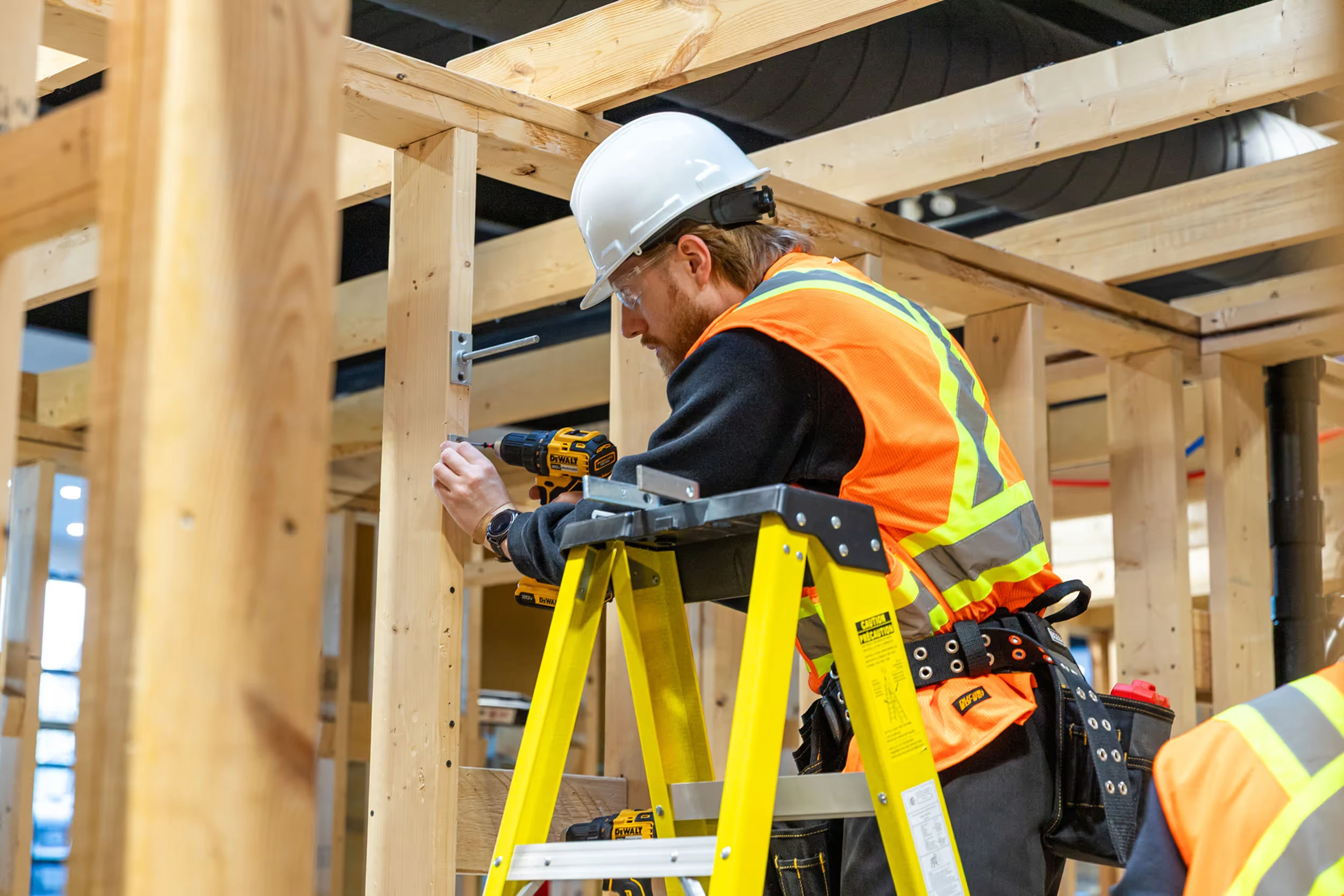 Construction worker wearing a white hard hat and orange safety vest using a power drill on wooden framing while standing on a yellow ladder inside a building under construction.