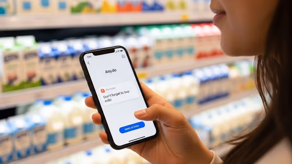 Close de mão segurando smartphone no supermercado com lembrete do app Any.do dizendo "Não esqueça de comprar leite".