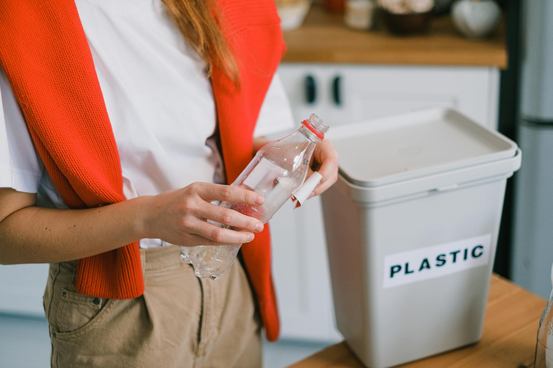 Person at home holding a poly mailer while standing next to curbside recycling bin and trash can, looking uncertain about proper disposal