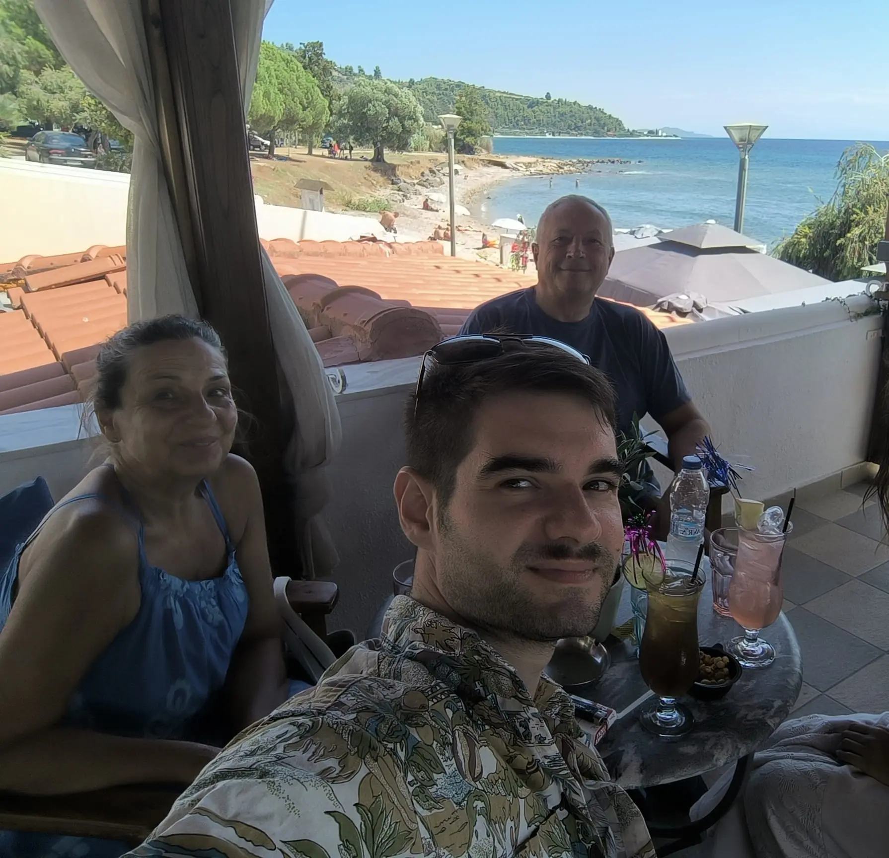 Three people sitting at a table with drinks on a terrace overlooking a beach and ocean under clear blue sky.