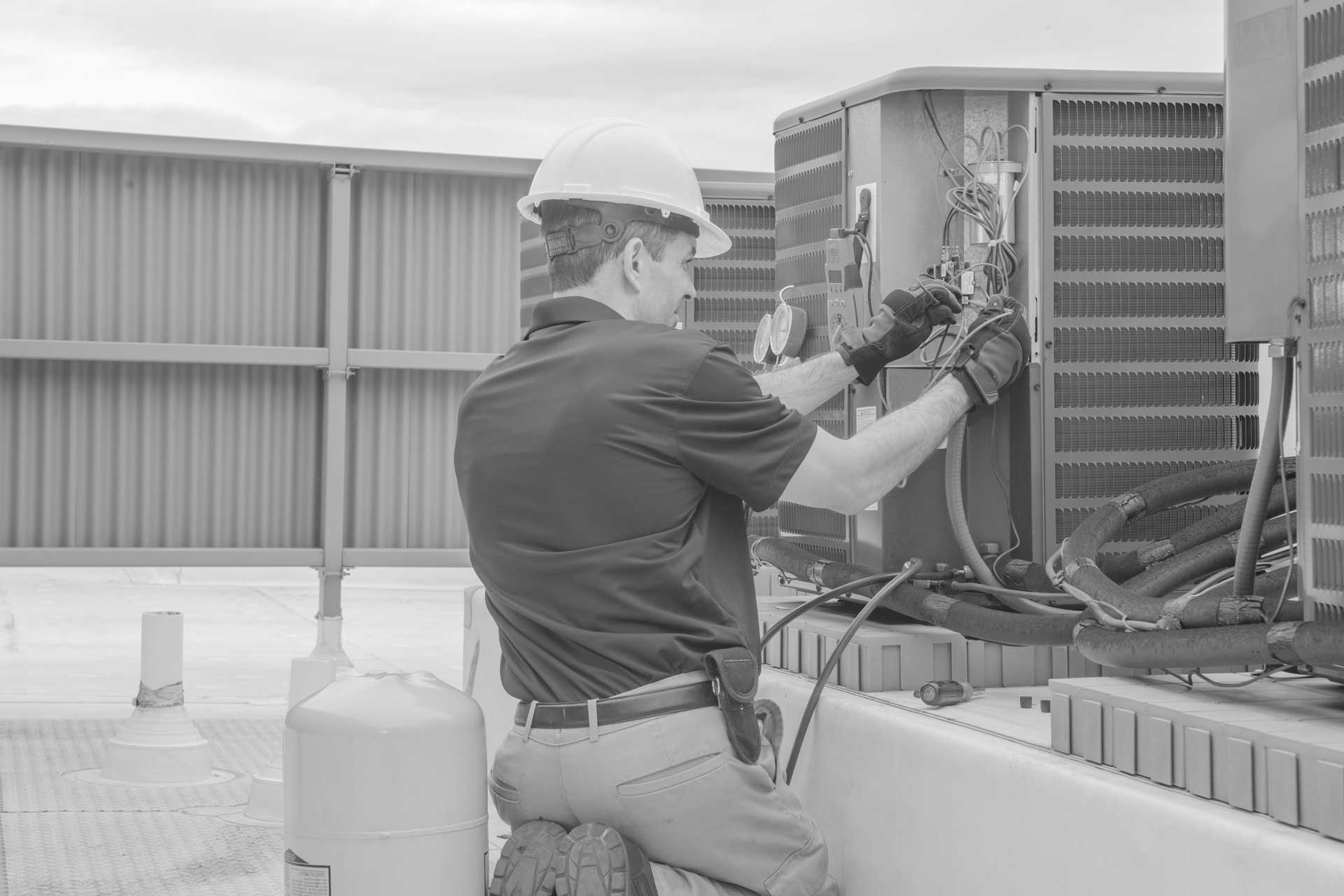 HVAC technician wearing a hard hat and gloves kneeling while inspecting and connecting wires on an outdoor air conditioning condenser unit.