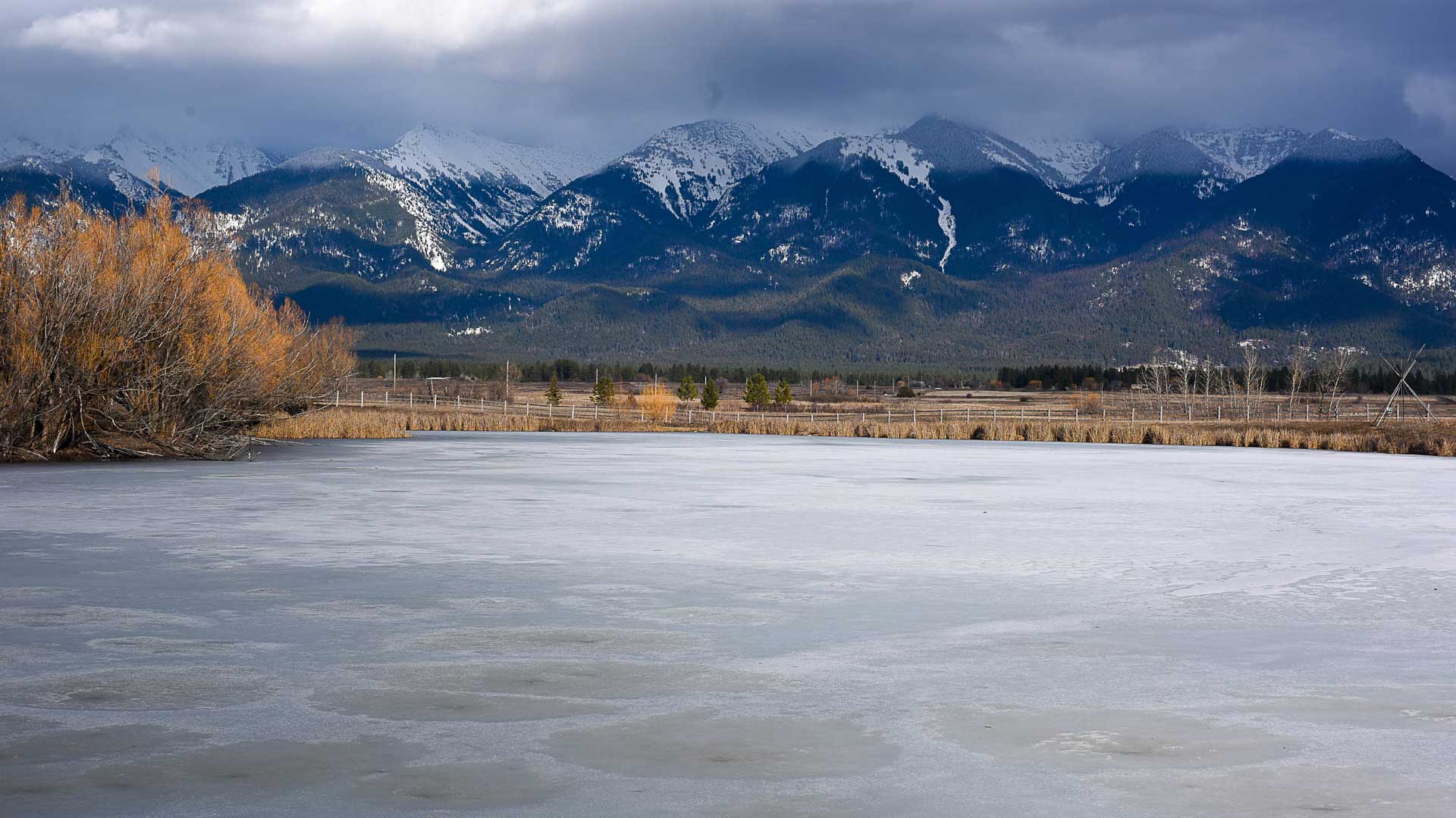 Frozen lake in front of leafless trees and a snow-capped mountain range under a cloudy sky.