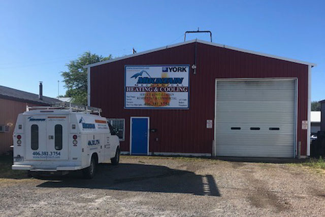 Red metal building with a large garage door and a blue entrance door, a white service van with ladder racks parked outside on a gravel lot under a clear blue sky.