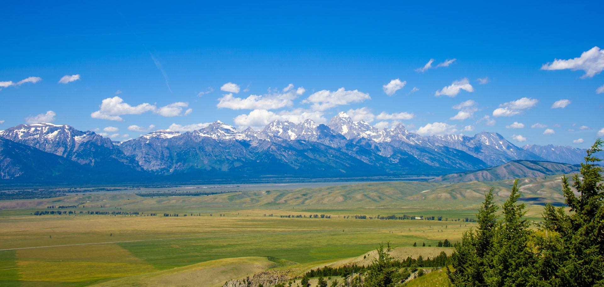 Wide view of a mountain range with snow-capped peaks under a blue sky with scattered clouds, overlooking a vast green valley with trees in the foreground.