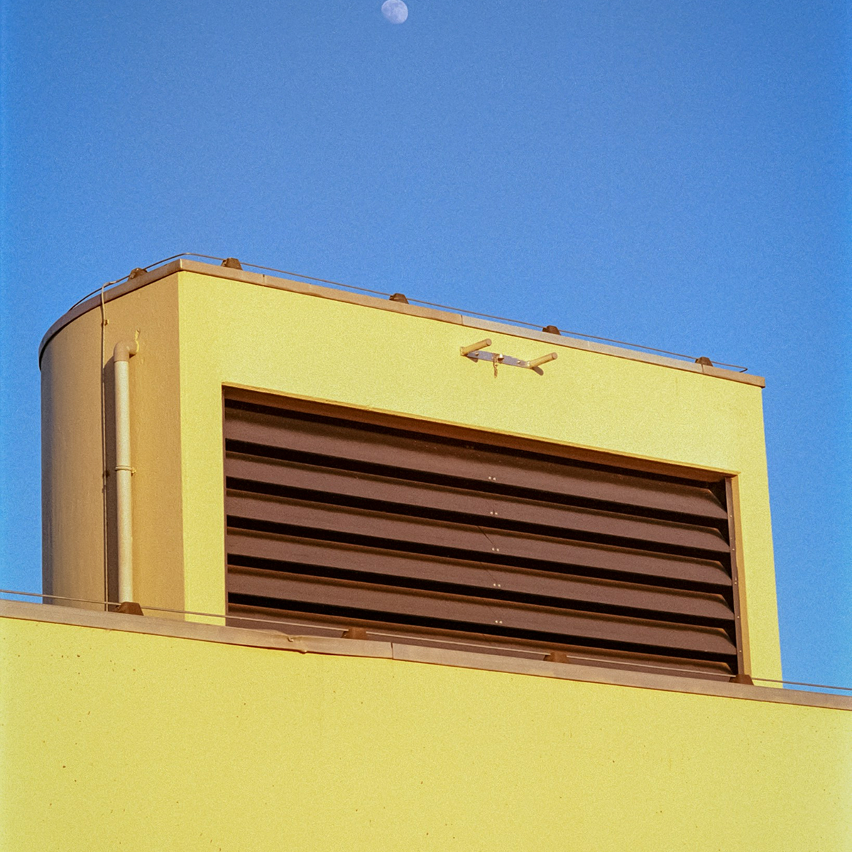 Yellow building rooftop structure with large dark louvered vent under a blue sky with a visible moon.