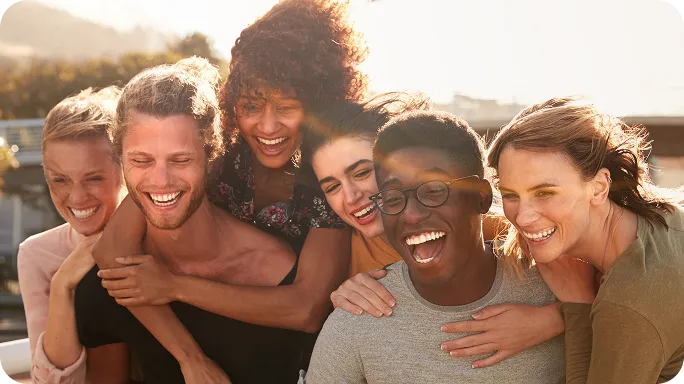 Group of six diverse friends laughing and embracing outdoors in bright sunlight.