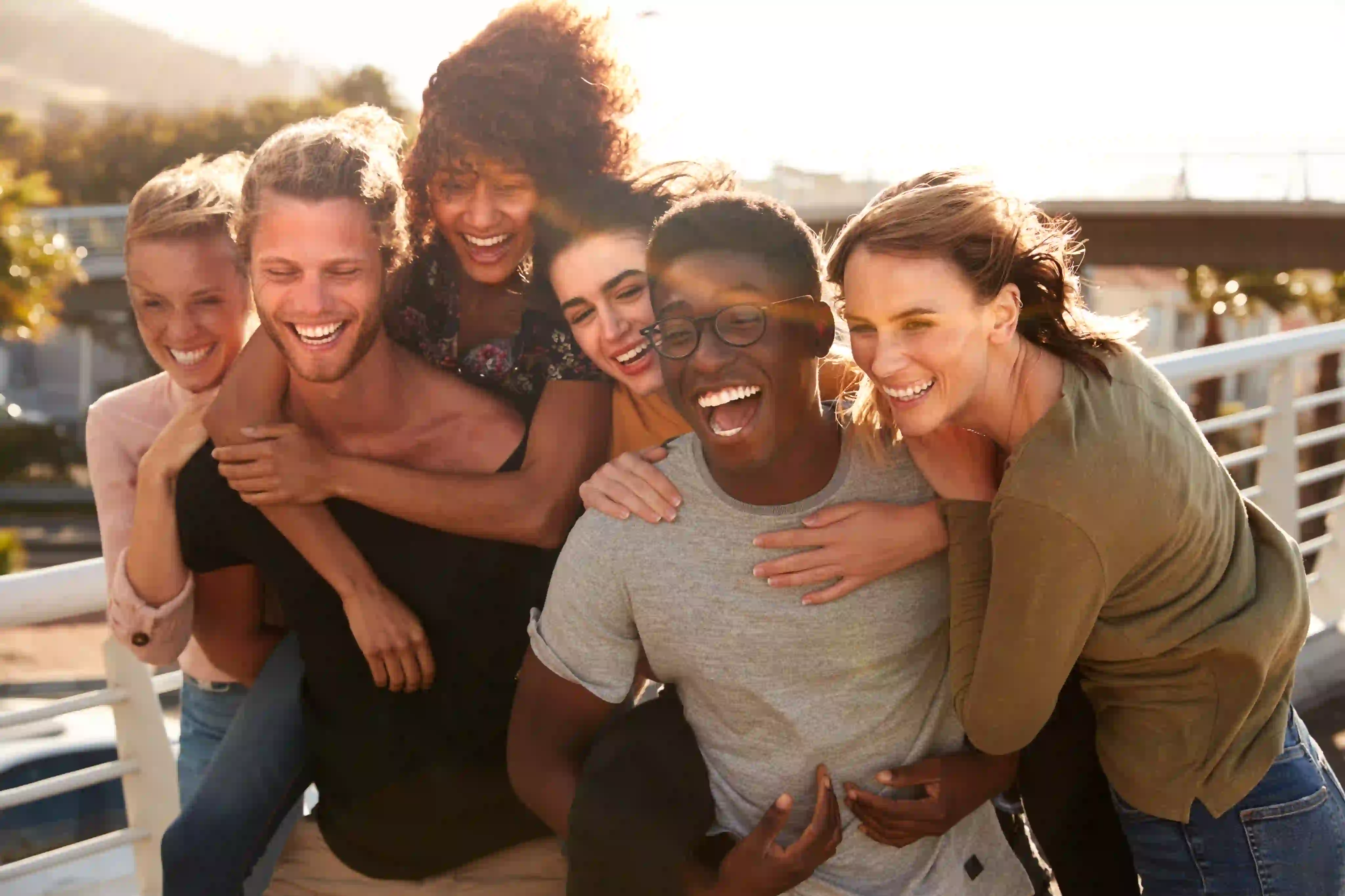 A group of six diverse friends smiling and laughing outdoors on a sunny day, with some giving piggyback rides.