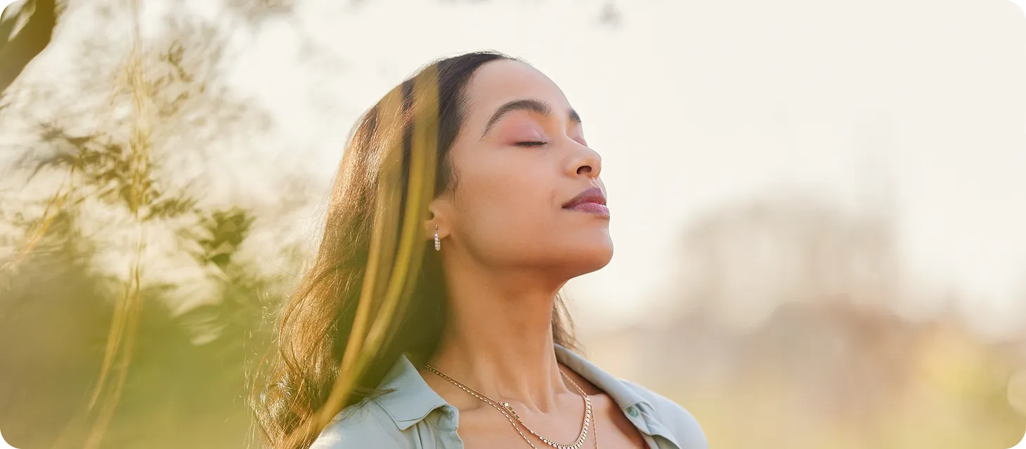 Woman with closed eyes enjoying sunlight outdoors amidst soft-focus greenery.