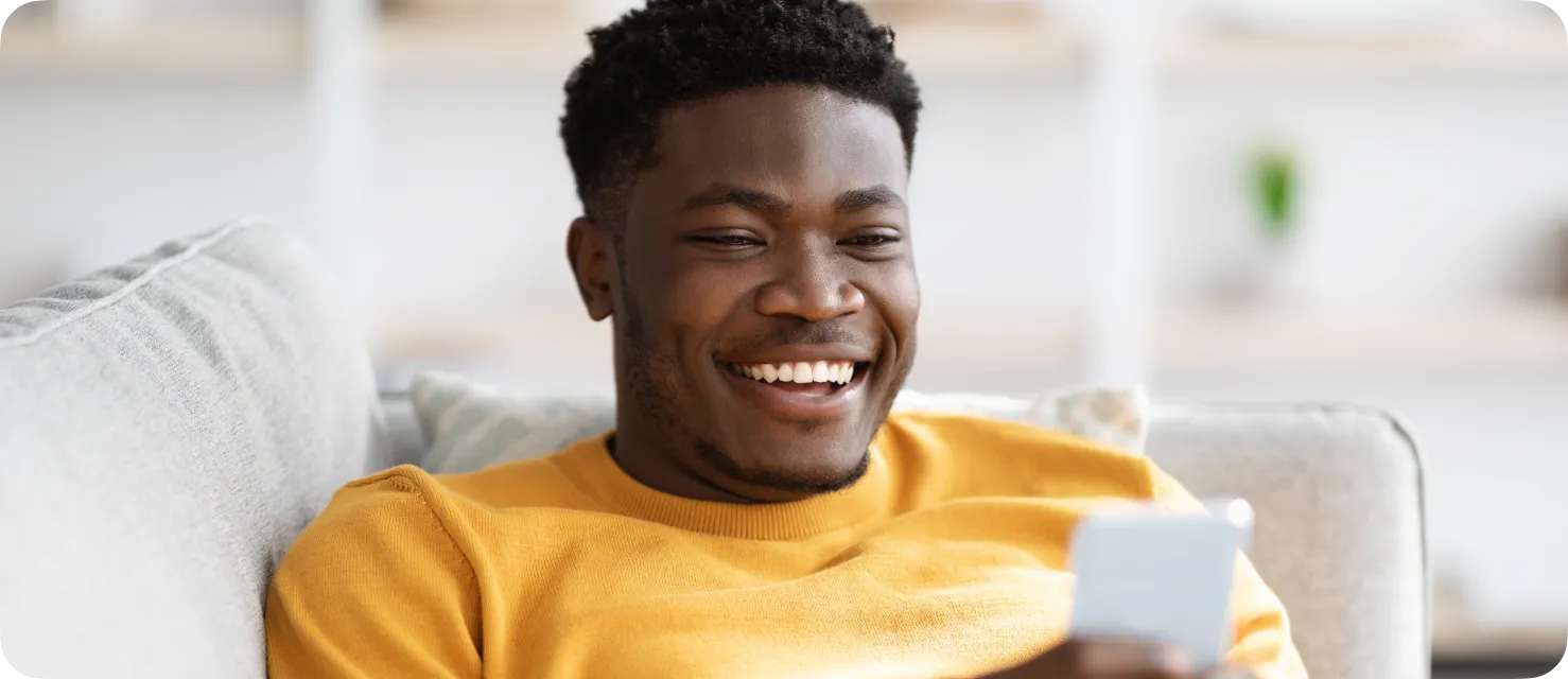Smiling young man in a yellow sweater looking at his smartphone while sitting on a light gray couch.