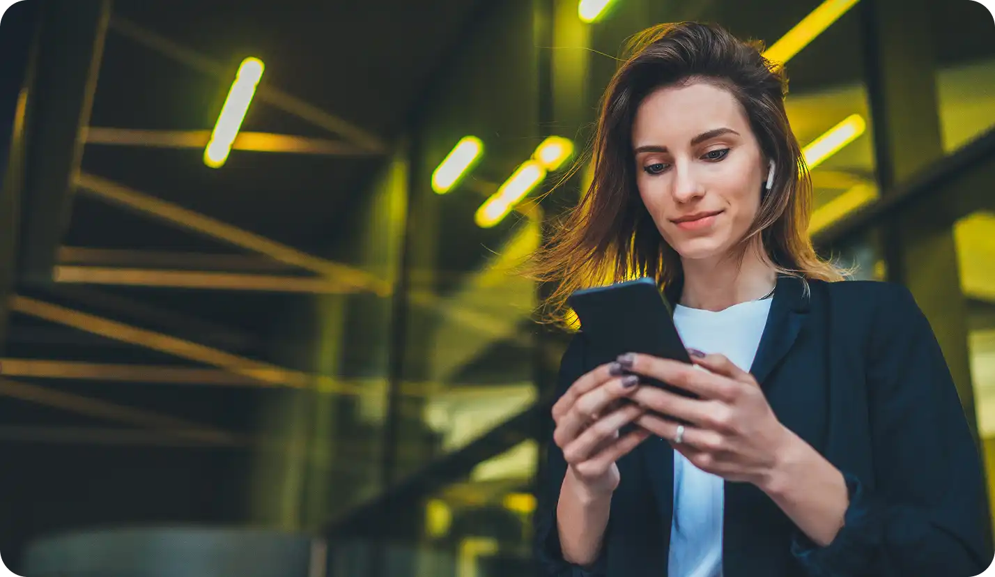 Young woman wearing wireless earbuds, looking at her smartphone with a slight smile in a modern indoor setting with yellow lights.