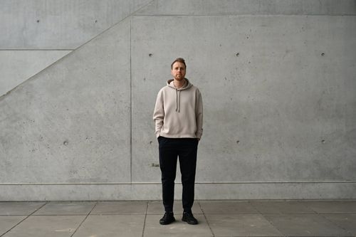 Oleksii Ananiev, oprichter van Storm Architects, staat voor een prachtige betonnen muur om een overzicht te geven van het uitzonderlijke afgelopen jaar van het bedrijf. In een informele stijl draagt hij een hoodie en broek.