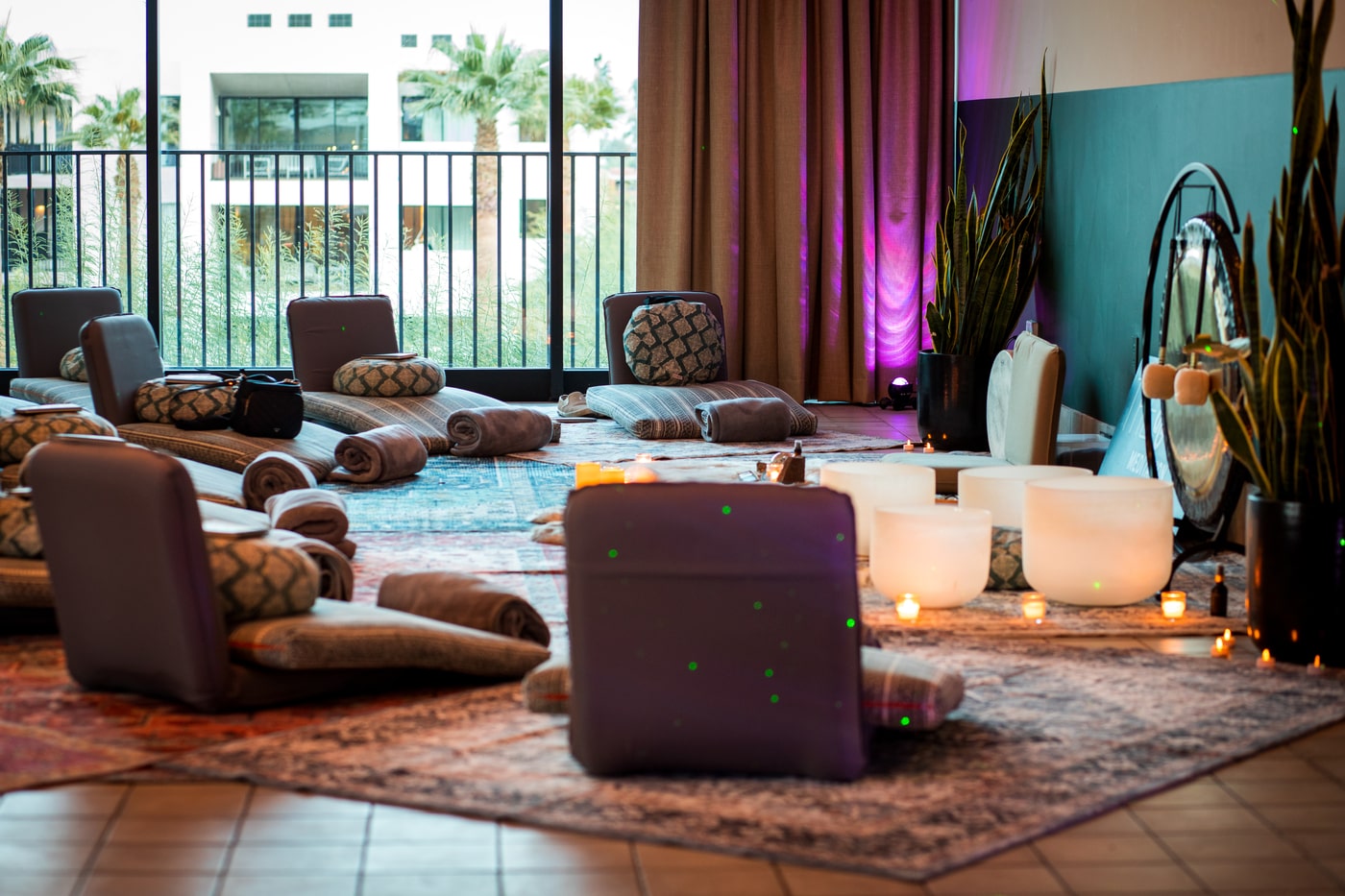 Cozy sound bath setup with floor cushions, candles, crystal bowls and gong by windows.