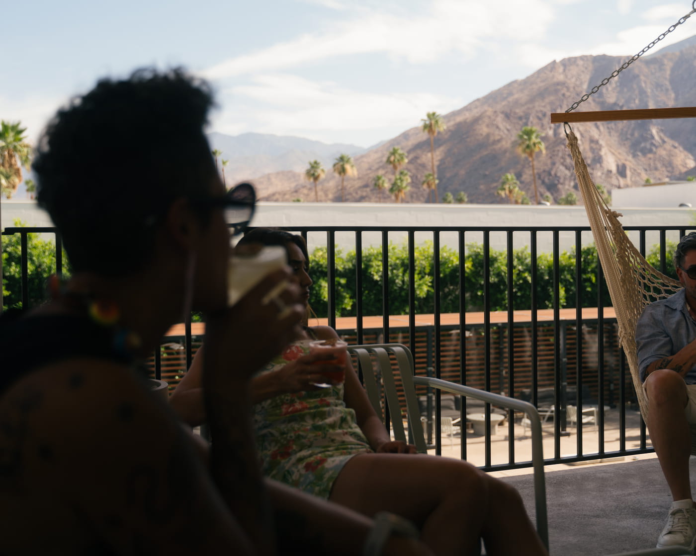 Friends relaxing on a balcony with drinks near a hammock, with palm trees and desert mountains in the background