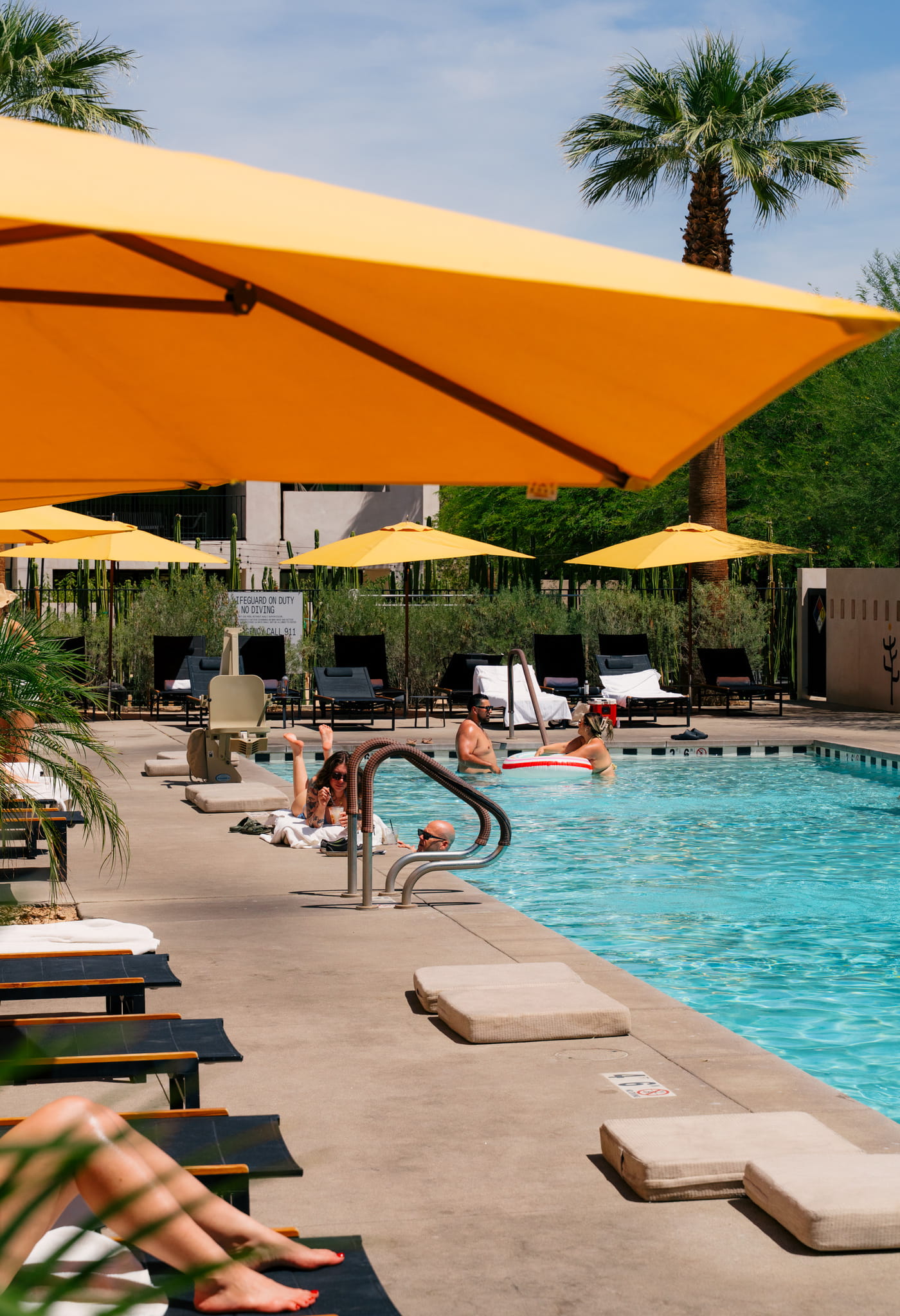 Guests lounging and swimming at a resort pool lined with yellow umbrellas, palm trees, and dark lounge chairs