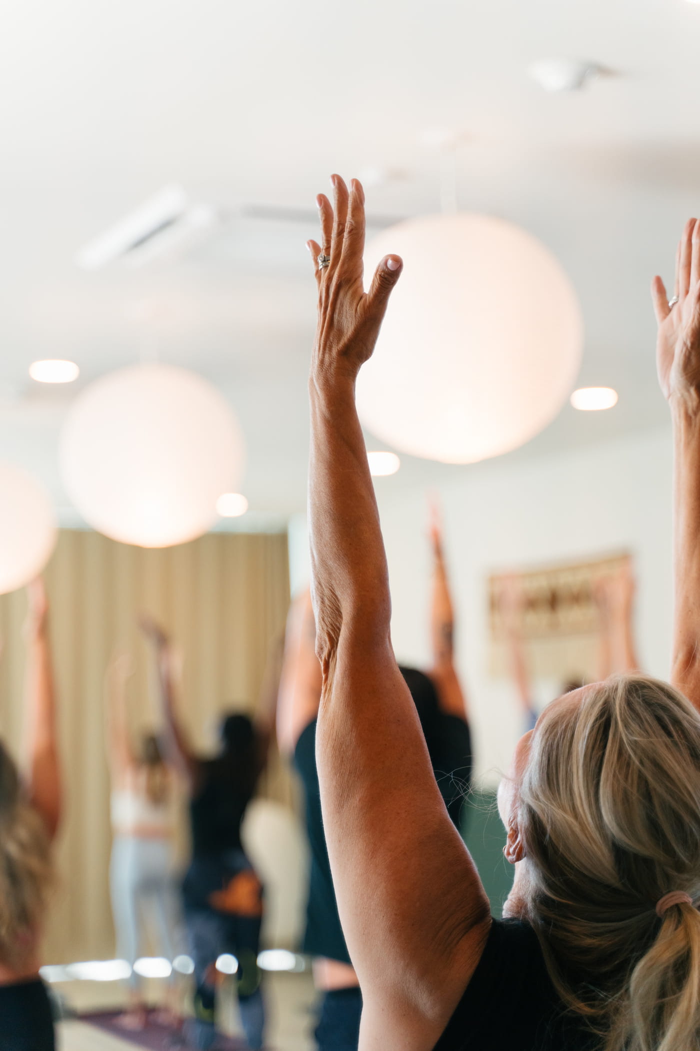 Participants raise their arms during a group yoga class in a bright, modern studio with round pendant lights.