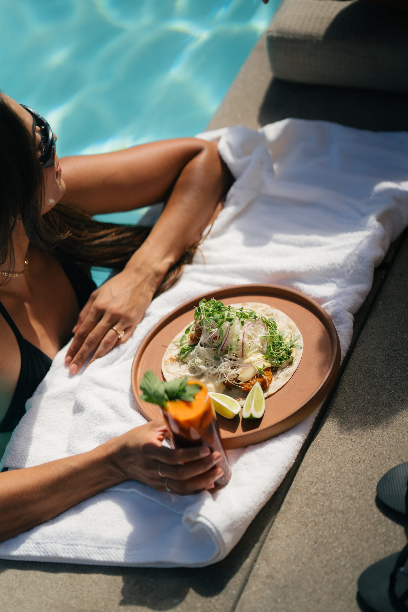 Woman lounging poolside in a black swimsuit with a gourmet fish taco plate and a cocktail on a white towel