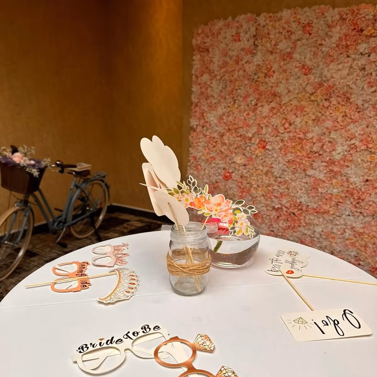 Round table with bridal shower photo props including glasses, rings, and floral decorations, with a pink flower wall and vintage bicycle in the background.