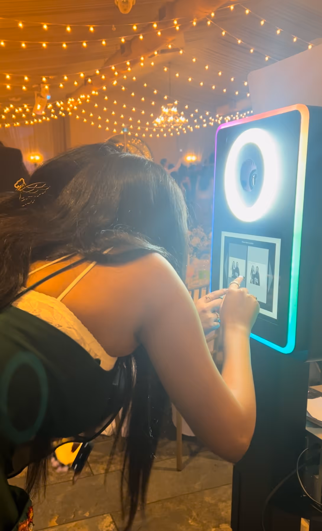 A woman using a digital photo booth touchscreen, adjusting photo options with her finger. The booth is illuminated with a bright circular ring light