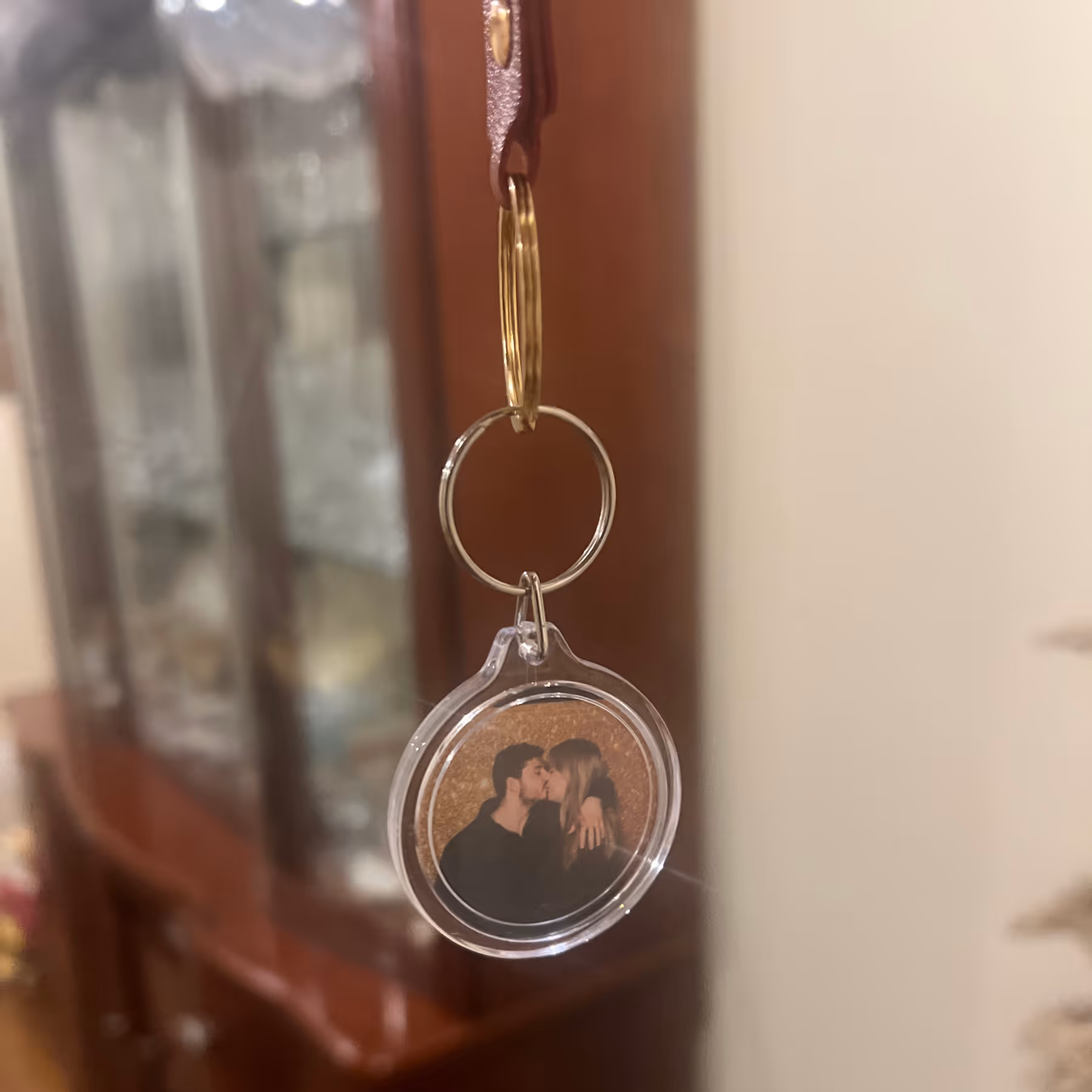 Keychain with a circular photo of a couple kissing, hanging inside a room with wooden furniture in the background.