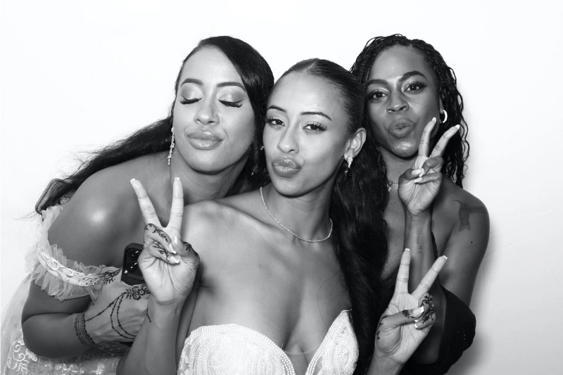 Three women posing closely together, each making peace signs with their fingers and puckering their lips, in a black and white photo.